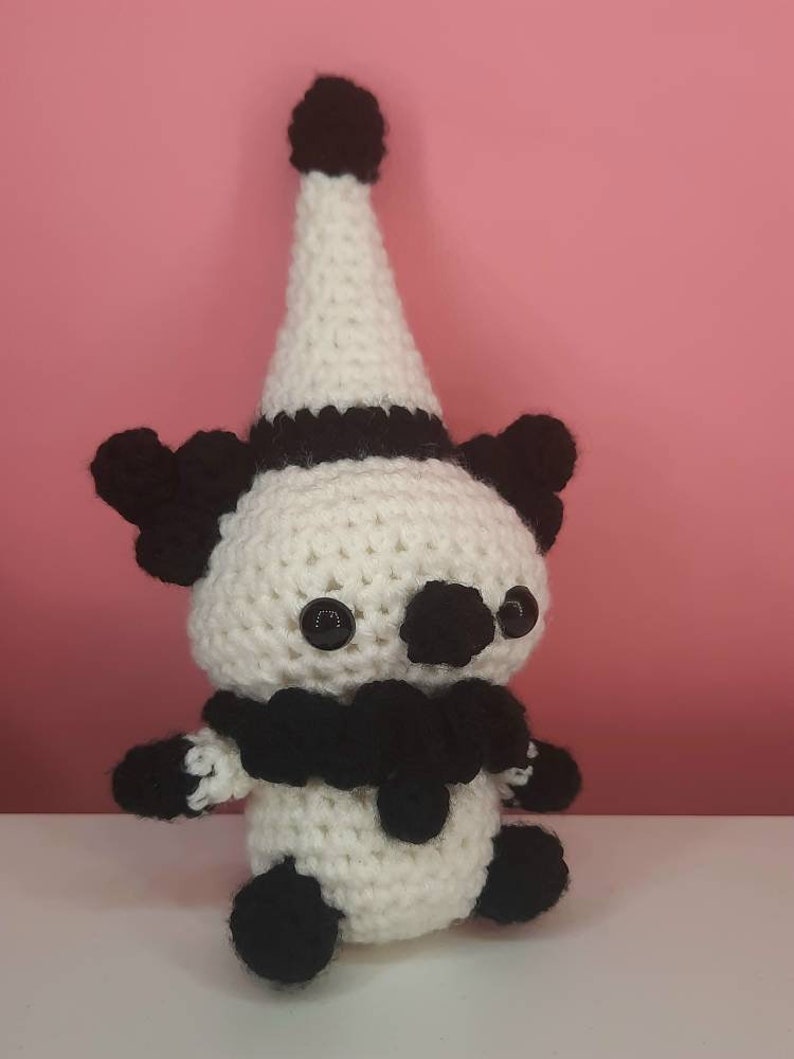 May include: A handmade crochet clown doll in white and black. The doll has a white cone hat with a black band and a black pom-pom on top. It has black button eyes, black ears, and a black ruffled collar. The doll's arms and feet are also black.