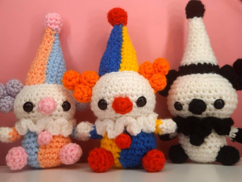 May include: Three handmade crochet clown dolls. The clowns have white bodies, colorful hats, and orange hair. One clown has a blue, yellow, and red hat, another has a blue, orange, and pink hat, and the third has a black and white hat.