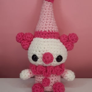 May include: A handmade crochet clown doll in pink and white. The clown has a white face, a pink nose, and large black eyes. It wears a pink ruffled collar, a pink and white hat, and has pink pom-poms on its body and ears. The doll is on a white surface.