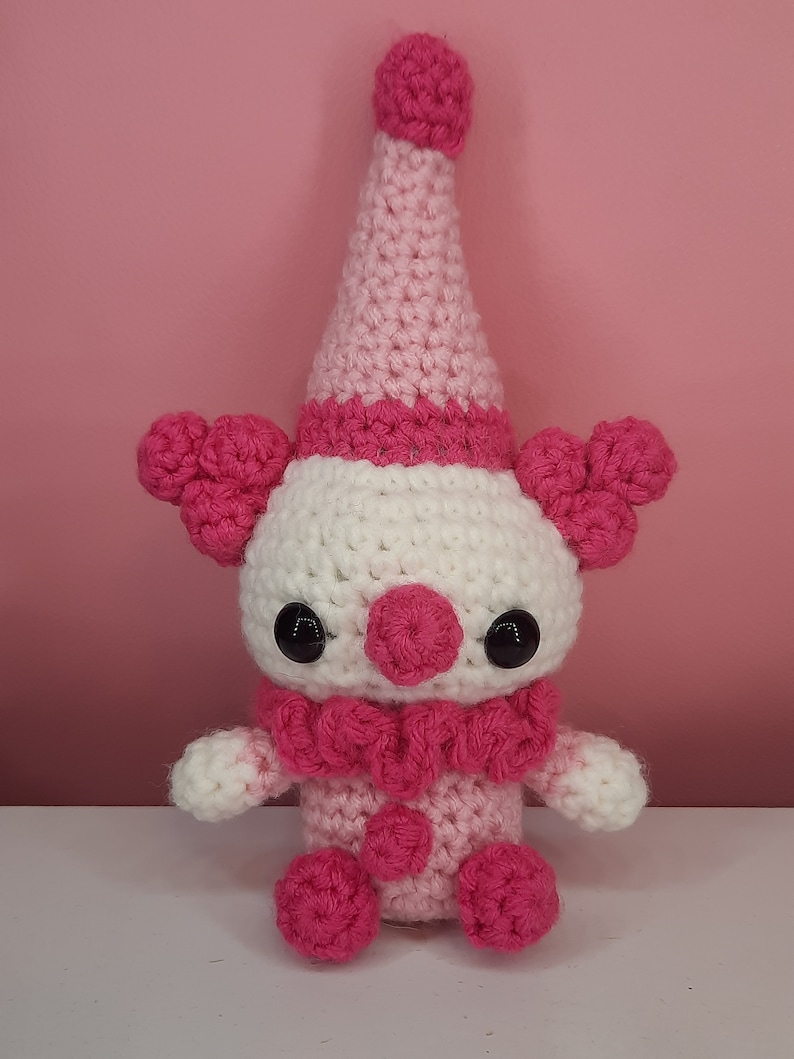 May include: A handmade crochet clown doll in pink and white. The clown has a white face, a pink nose, and large black eyes. It wears a pink ruffled collar, a pink and white hat, and has pink pom-poms on its body and ears. The doll is on a white surface.