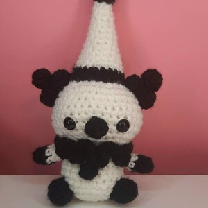 May include: A handmade crochet doll in a clown-like design. The doll features a white body, black accents, and a tall, pointed hat. The doll has black eyes, a black scarf, and small black pom-poms on the hat and ears.