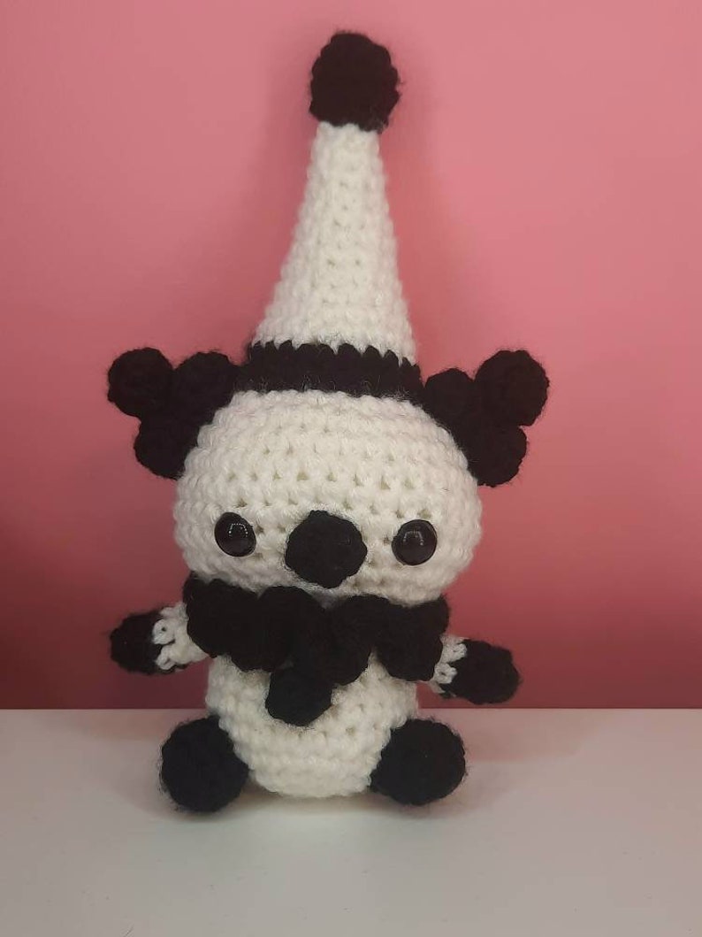 May include: A handmade crochet doll in a clown-like design. The doll features a white body, black accents, and a tall, pointed hat. The doll has black eyes, a black scarf, and small black pom-poms on the hat and ears.