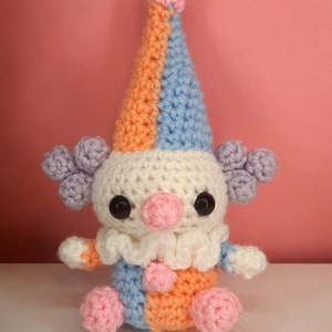 May include: A handmade crochet clown doll with a colorful design. The clown features a tall, pointed hat in orange, blue, and pink, with matching accents on the body. It has large black eyes, a pink nose, and a white ruffled collar.