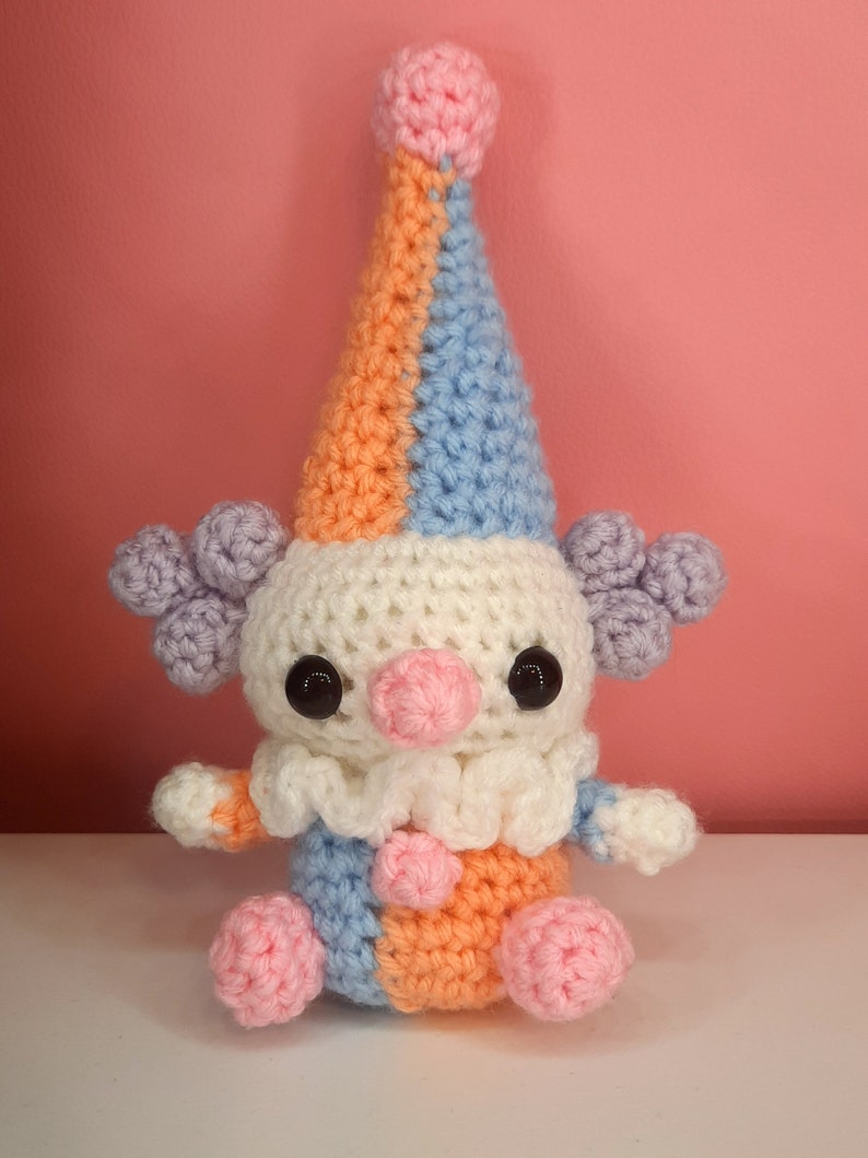 May include: A handmade crochet clown doll with a colorful design. The clown features a tall, pointed hat in orange, blue, and pink, with matching accents on the body. It has large black eyes, a pink nose, and a white ruffled collar.