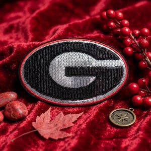 May include: An oval-shaped patch featuring a large letter G in black and silver sequins. The design is outlined with a thin red border. The patch is displayed alongside red berries, a dried maple leaf, and a small circular metal compass.
