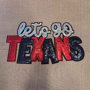 May include: A decorative patch featuring the text "lets go" in iridescent silver sequins above the word "TEXANS" in large, alternating red and black sequins. The letters are outlined in black, creating a bold, high-contrast design.