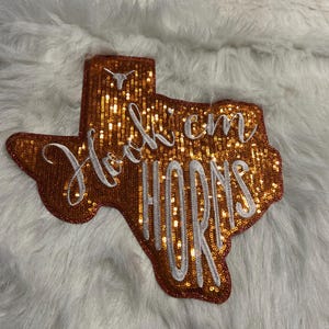 May include: A Texas-shaped patch covered in shimmering orange sequins. The patch features white embroidered text that reads Hook 'em HORNS in a mix of cursive and block lettering, with a small white longhorn steer logo at the top center.