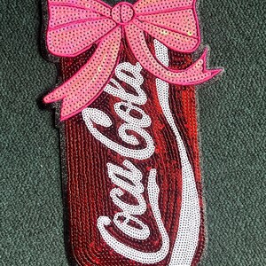 May include: A red and white Coca-Cola can with a pink sequined bow on top. The can is made of sequins and the bow is made of iridescent sequins.