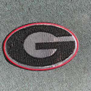 May include: A black and silver sequined patch with a red outline, featuring a large letter 'G' in the center.