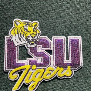 May include: A sequined patch featuring the LSU Tigers logo. The design includes a yellow tiger head, the letters "LSU" in purple, and the word "Tigers" in yellow. The patch is outlined in white.