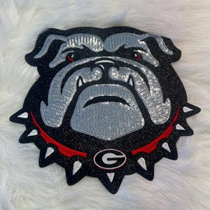May include: A black and silver sequined bulldog head patch with a red and white collar and a black and white 'G' in the center.