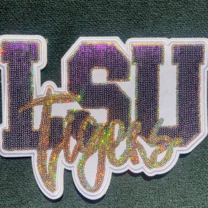 May include: A white patch with the letters "LSU" in purple, sequined letters. The word "Tigers" is written in a shimmering, gold sequined script across the bottom of the patch. The patch has a white border.