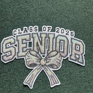 May include: A white and iridescent sequined patch with the words "CLASS OF 2026 SENIOR" in a bold, outlined font. A sequined bow is centered below the word "SENIOR". The patch is on a green textured background.