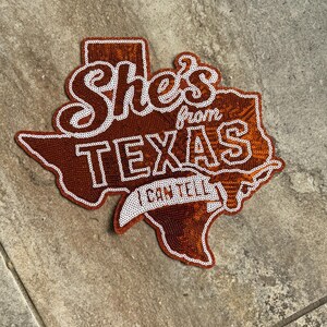 May include: A Texas-shaped patch covered in burnt orange sequins with white embroidered borders and text. The patch features the words She's from TEXAS I CAN TELL in white script and block lettering, along with a small white longhorn steer icon.