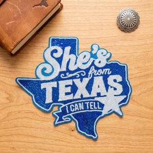 May include: A blue and white beaded patch shaped like the state of Texas. The patch features the text "She's from TEXAS I CAN TELL" in white beads against a blue beaded background, with a white beaded star accent on the right side of the design.