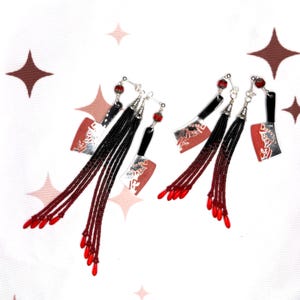 May include: A pair of novelty earrings featuring a cleaver design. The earrings have a silver blade with red accents, a black handle, and black and red beaded tassels. The top of the earrings have a red bead.