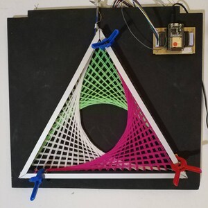 Könnte beinhalten: Ein weißer dreieckiger Rahmen mit grünen, weißen und rosa Fäden, die darüber gespannt sind und ein geometrisches Muster bilden. Der Rahmen ist mit blauen und roten Klemmen an einer schwarzen Oberfläche befestigt. In der oberen rechten Ecke ist eine kleine Platine mit Drähten zu sehen.