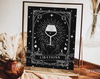 May include: Black and white art print in a gold frame. The print features a wine glass with a floral design, radiating lines, and the word "LIBATIONS" on a banner. The design includes stars, moons, and hands.