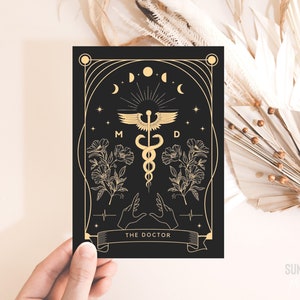 Puede incluir: Impresión artística en negro y dorado con un símbolo médico alado, un caduceo y el texto "THE DOCTOR". El diseño incluye elementos florales, lunas crecientes y las letras "M" y "D".