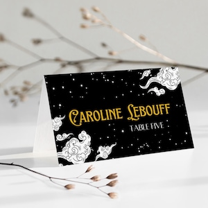 May include: A folded place card with a black background featuring white clouds and stars. The text "Caroline Lebouff" is in gold script, with "Table Five" below. The card is on a white surface.