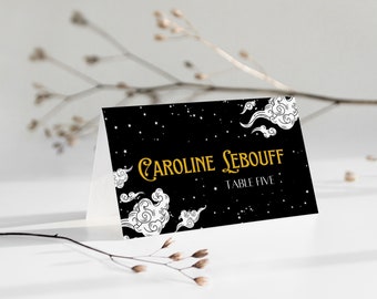May include: A folded place card with a black background featuring white clouds and stars. The text "Caroline Lebouff" is in gold script, with "Table Five" below. The card is on a white surface.
