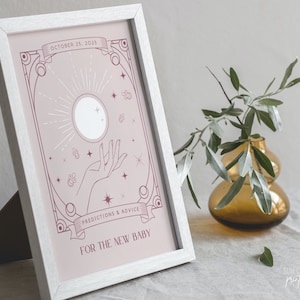 May include: A framed print with a light pink background features a celestial design with a hand, sun, and stars. Text reads "OCTOBER 25, 2025" and "PREDICTIONS & ADVICE FOR THE NEW BABY". The frame is white.
