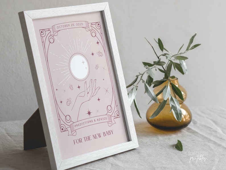 May include: A framed print with a light pink background features a celestial design with a hand, sun, and stars. Text reads "OCTOBER 25, 2025" and "PREDICTIONS & ADVICE FOR THE NEW BABY". The frame is white.