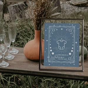 May include: A framed blue sign with white text and a onesie graphic, reading "Decorate a Onesie for the Baby." The sign is on a wooden table with champagne glasses and a terracotta vase. The background is blurred.
