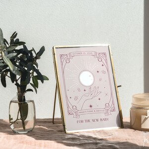 May include: A framed print with a gold-colored stand features a light purple design with the text "October 25, 2025", "Predictions & Advice", and "For The New Baby". A small glass vase with green leaves and a candle are also in the image.