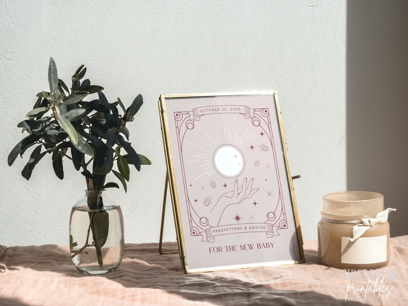 May include: A framed print with a gold-colored stand features a light purple design with the text "October 25, 2025", "Predictions & Advice", and "For The New Baby". A small glass vase with green leaves and a candle are also in the image.