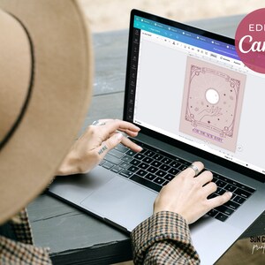 May include: A laptop displaying a digital design with a celestial theme, featuring a sun, hand, and stars, with the text "EDIT IN Canva" in a pink circle. The design includes the text "FOR THE NEW BABY".