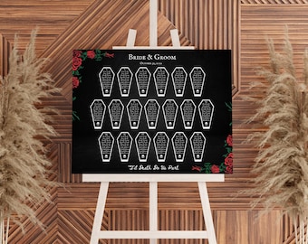 May include: A black seating chart with coffin-shaped table assignments, red roses, and the text "Bride & Groom" and "'Til Death Do Us Part." The chart is displayed on a white easel against a wooden backdrop.