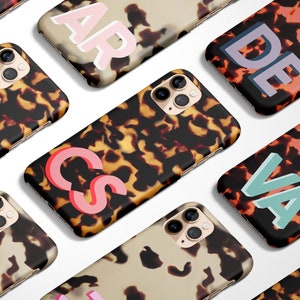May include: A collection of iPhone cases with a tortoiseshell pattern and colorful, oversized initials. The cases are arranged in a staggered pattern on a white background.