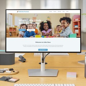 May include: A computer monitor displays a website for Little Stars Preschool, with the tagline "Where Little Minds Grow Big Dreams." The monitor sits on a wooden desk with a lamp, plant, and other office supplies.