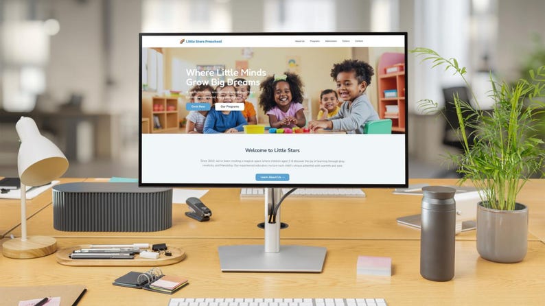 May include: A computer monitor displays a website for Little Stars Preschool, with the tagline "Where Little Minds Grow Big Dreams." The monitor sits on a wooden desk with a lamp, plant, and other office supplies.