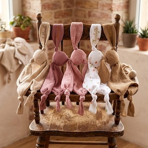 Può includere: Una collezione di copertine di sicurezza per bambini in vari colori, tra cui beige, rosa e bianco, è esposta su una sedia di legno. Ogni copertina presenta una testa annodata e un'etichetta con un nome ricamato. Le copertine sono realizzate in tessuto morbido e strutturato.