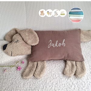 May include: A rectangular brown pillow shaped like a dog with floppy ears and a tail. The centre of the pillow features the name Jakob embroidered in white script. Above the pillow are four small circular icons showing a bear, sheep, goose, and grass.