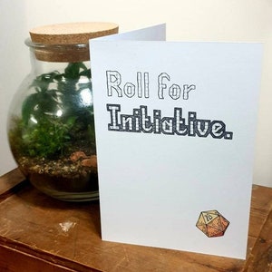 May include: A white card with black text that reads "Roll for Initiative." A cartoon drawing of a 20-sided die is in the bottom right corner of the card.