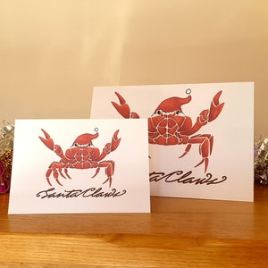 May include: Two white greeting cards featuring red crabs wearing Santa hats. The cards are inscribed with the text "Santa Claws".