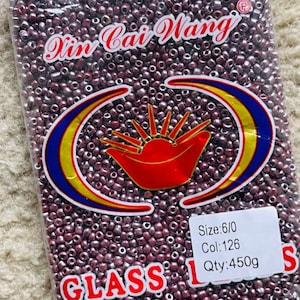 May include: A transparent bag filled with small, dark red glass beads. The bag features the text "Xin Cai Wang" and a graphic. The label also indicates the size, colour, and quantity of the beads, which is 450g.