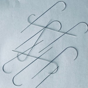 Puede incluir: Primer plano de un grupo de ganchos de metal de color plateado, algunos doblados en forma de gancho y otros rectos. Los ganchos est&aacute;n dispuestos en un patr&oacute;n aleatorio sobre una superficie azul claro.