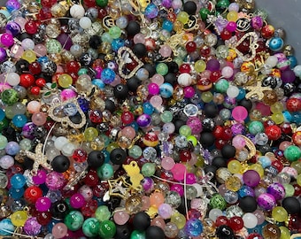 May include: A container overflowing with a mix of colourful beads and charms. The beads are diverse in size, shape, and hue, including red, pink, green, and black. Charms feature butterflies, hearts, and stars, creating a vibrant assortment.