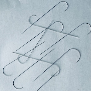 Puede incluir: Primer plano de un grupo de ganchos de metal de color plateado, algunos doblados en forma de gancho y otros rectos. Los ganchos est&aacute;n dispuestos en un patr&oacute;n aleatorio sobre una superficie azul claro.