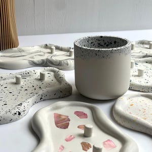 May include: A collection of decorative trays and a small ceramic cup. The trays are in various shapes and patterns, including terrazzo and marble designs. The cup is white with a speckled rim. The trays have small, cylindrical pegs.