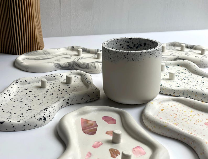 May include: A collection of decorative trays and a small ceramic cup. The trays are in various shapes and patterns, including terrazzo and marble designs. The cup is white with a speckled rim. The trays have small, cylindrical pegs.