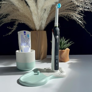 Puede incluir: Un cepillo de dientes eléctrico con un cabezal azul y blanco descansa sobre una bandeja moteada de color verde menta. Un tubo de pasta de dientes está en una taza bicolor verde menta y blanca. Hierba de la pampa decorativa y una suculenta en jarrones de madera.