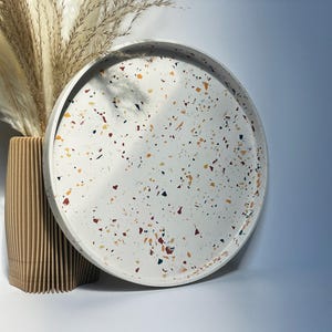 May include: A circular, white terrazzo tray with colourful flecks of red, yellow, and black. Beside it is a ribbed, light brown vase holding dried pampas grass. The background fades from white to light blue.