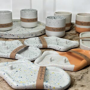 May include: A collection of speckled ceramic candle holders and trays. The candle holders are cylindrical, with a white base and speckled gray top. The trays are white with colorful speckles, some with a peach accent. Each item has a brown paper band that says "Cozy Candle Barn Co."