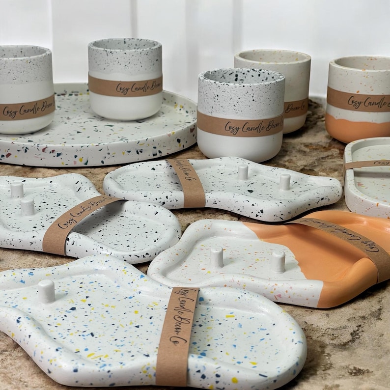 May include: A collection of speckled ceramic candle holders and trays. The candle holders are cylindrical, with a white base and speckled gray top. The trays are white with colorful speckles, some with a peach accent. Each item has a brown paper band that says "Cozy Candle Barn Co."