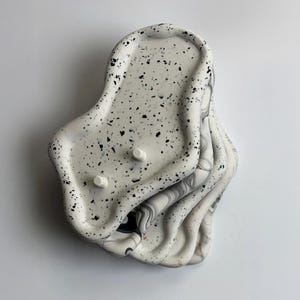 May include: A stack of four irregularly shaped, speckled white trays with black flecks. The top tray has two small, white, circular protrusions. The edges of the trays have a wavy design, and the bottom trays have a marbled pattern.